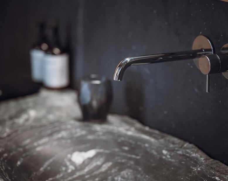 Detailansicht von einem Steinwaschbecken mit Pflegeprodukten und dunkeln Fliesen.
